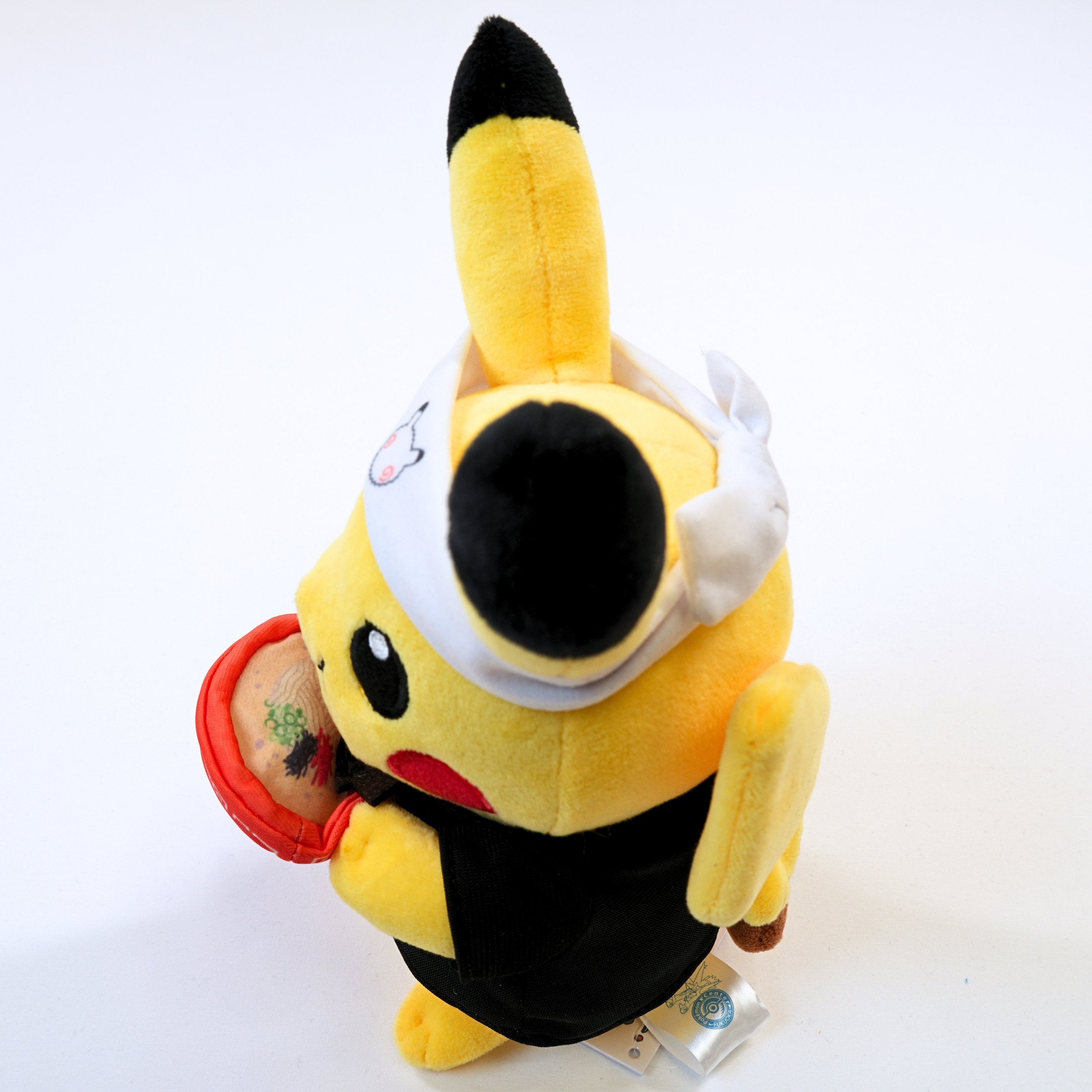 Vista lateral del peluche Pokémon Center Fukuoka R Ramen Pikachu, mostrando el volumen del peluche, la ropa y el cuenco